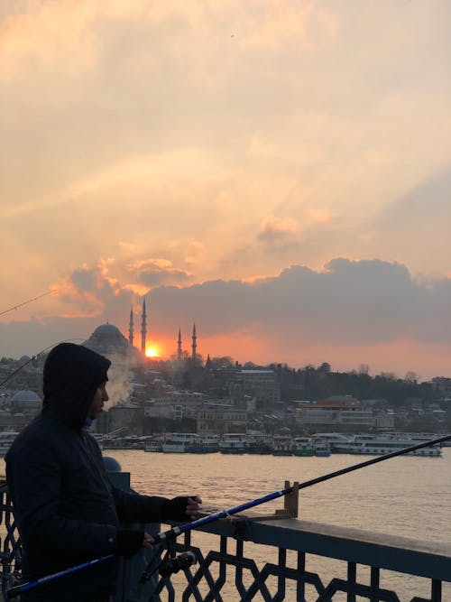 Man in Hoodie Jacket Standing Near the Metal Railing while Fishing During Sunset
