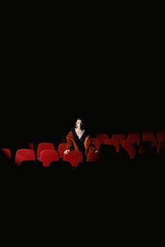 A solitary woman in a dimly lit cinema surrounded by empty red seats.