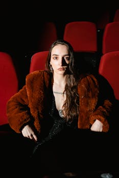 A woman with long hair sits in a cinema wearing a fur coat, exuding a serious expression.