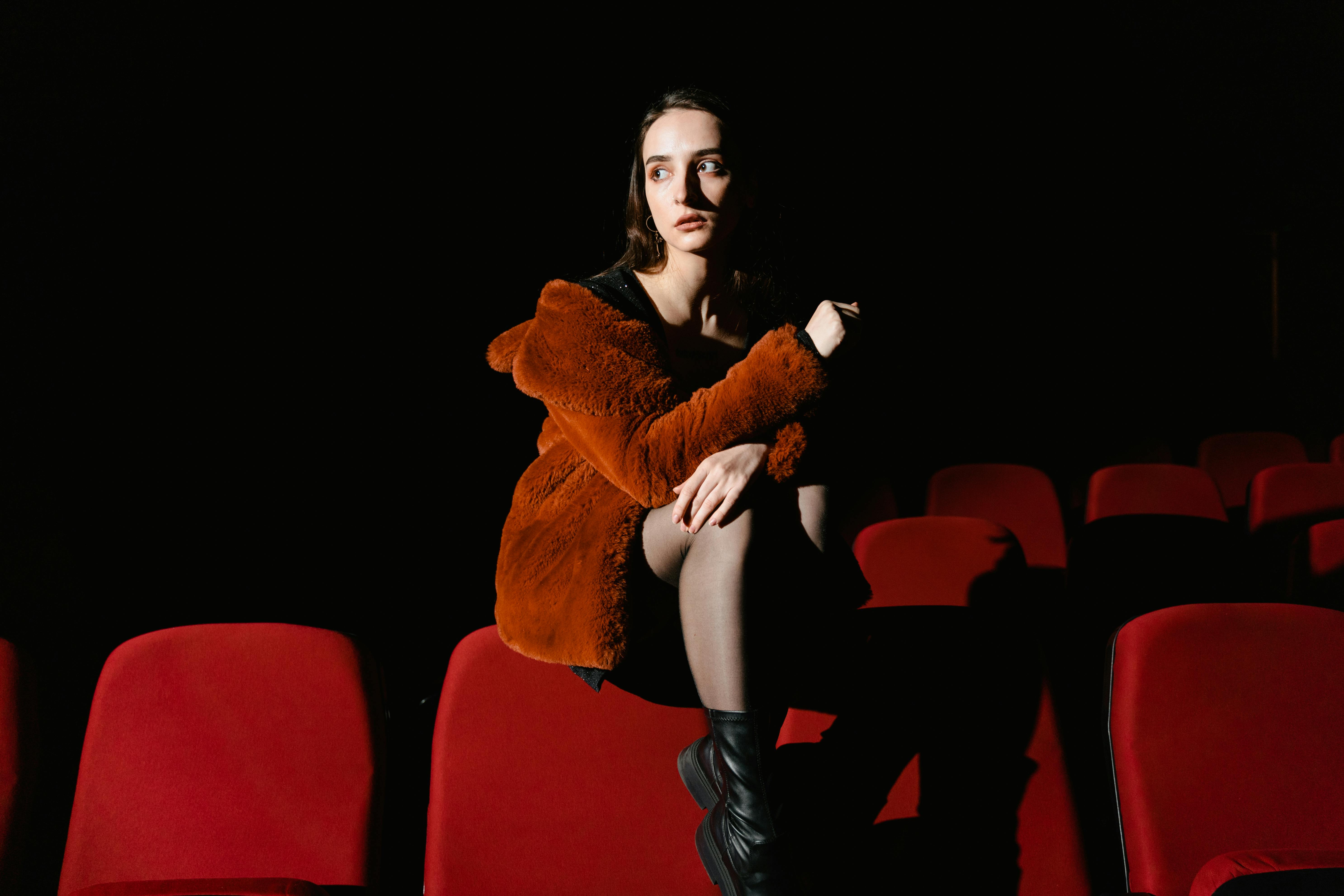 Free A woman in a fur jacket sits in a dimly lit movie theater with red seats. Stock Photo