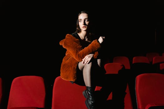 A woman in a fur jacket sits in a dimly lit movie theater with red seats.