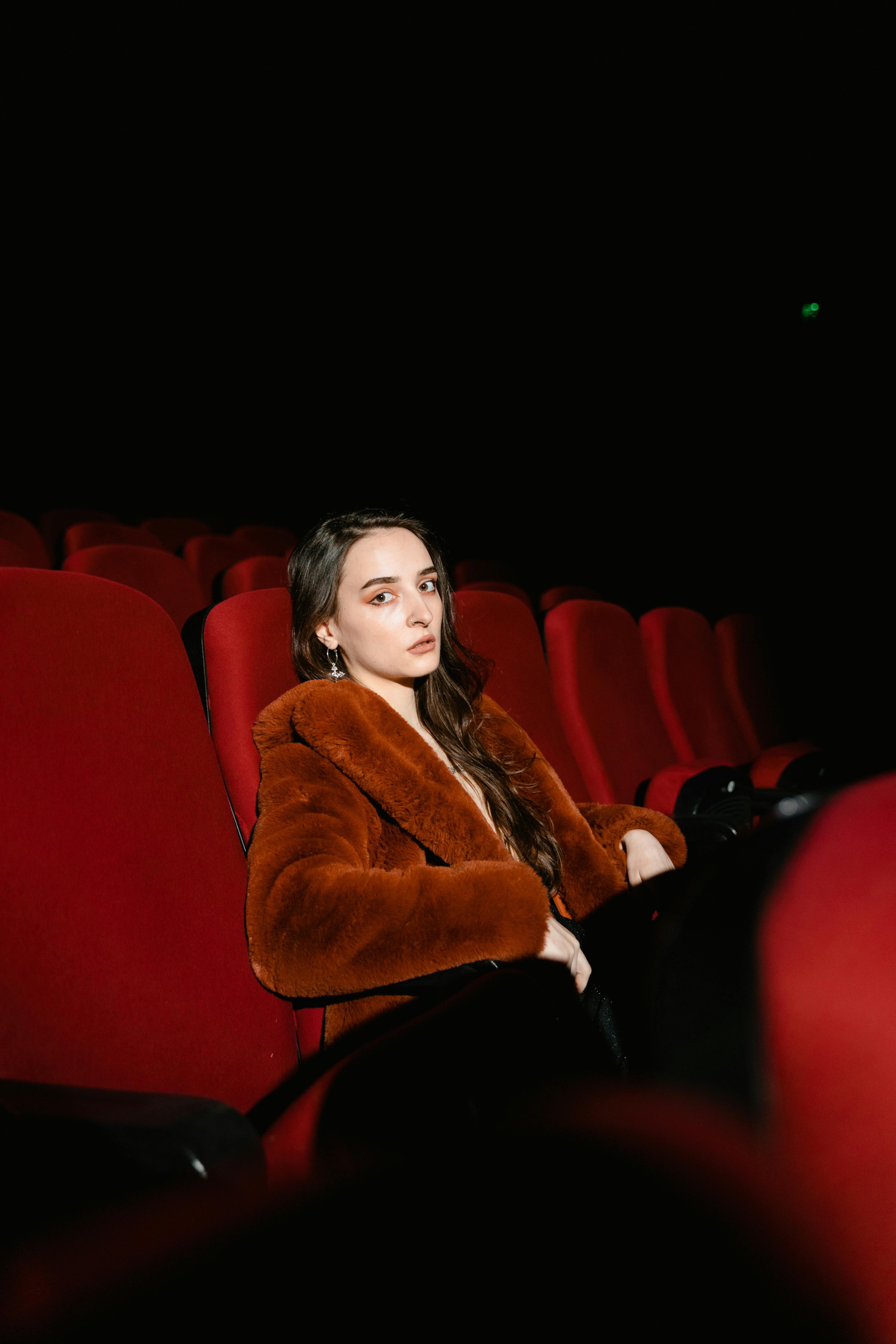 Free Young woman sitting alone in an empty cinema wearing a brown coat, surrounded by red seats. Stock Photo