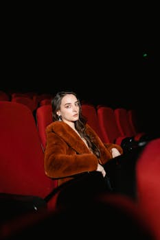 Young woman sitting alone in an empty cinema wearing a brown coat, surrounded by red seats.