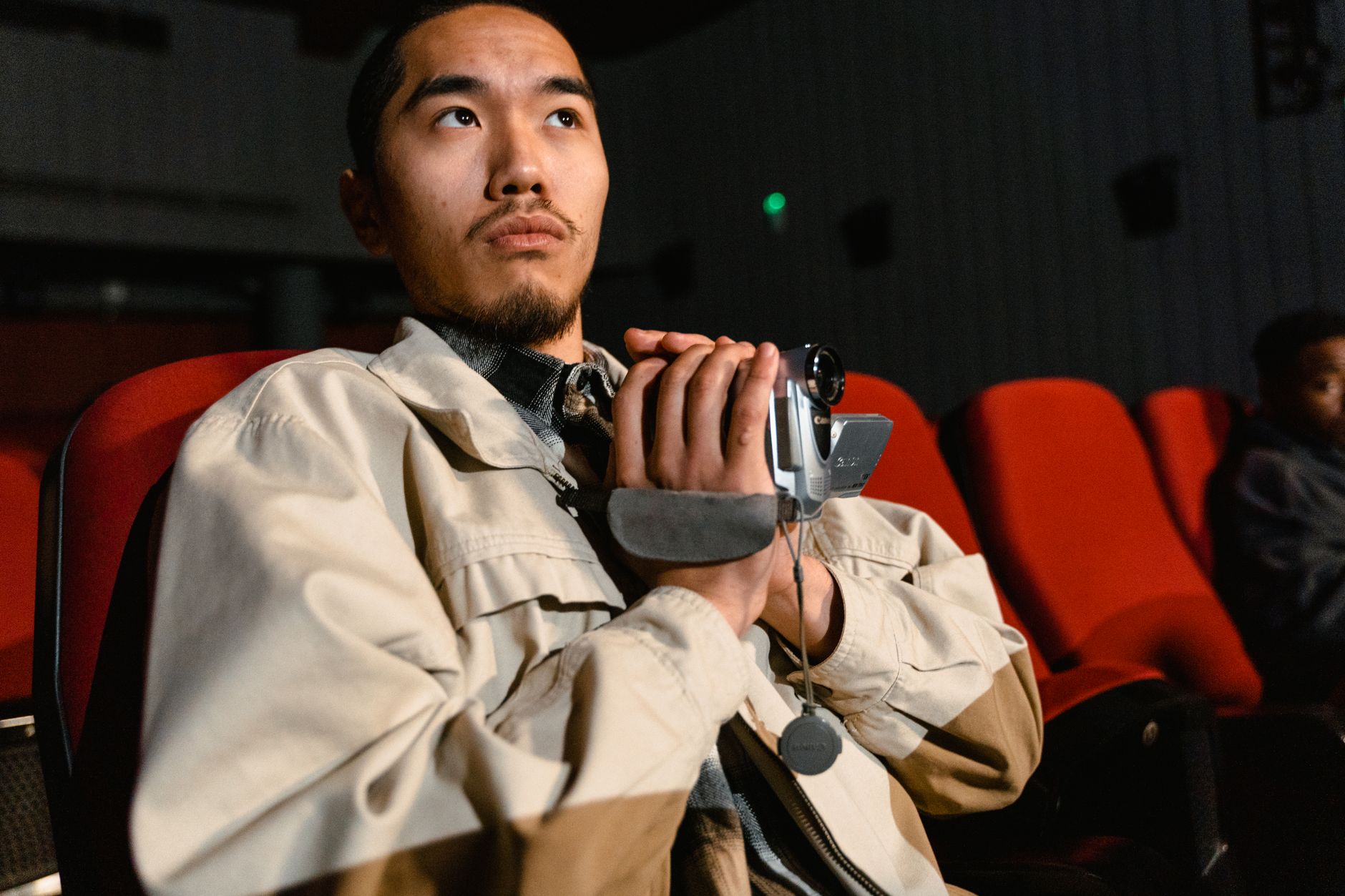 An adult male sitting in a cinema, holding a video camera, with red seats around.