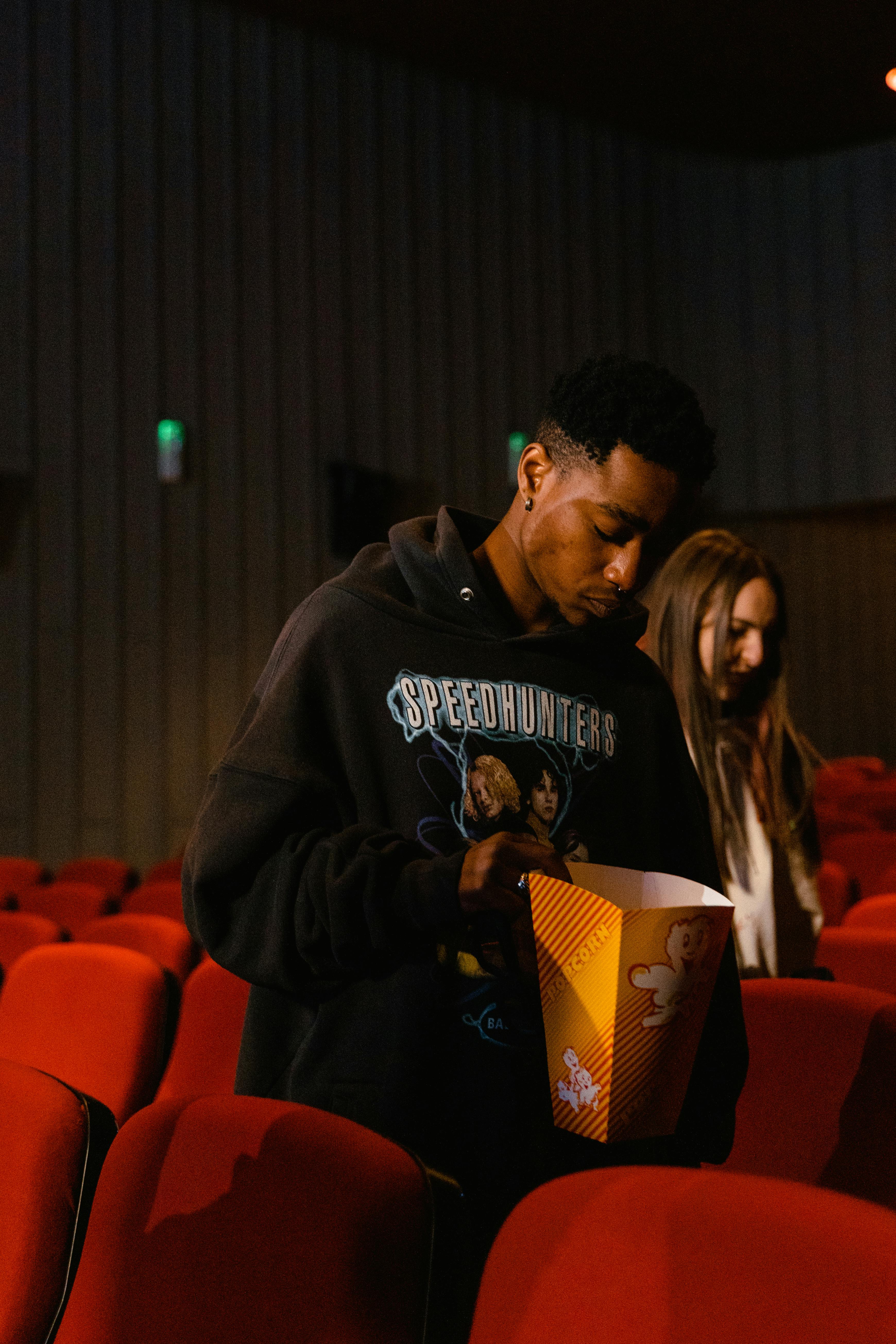 Free A man in a black hoodie holding popcorn inside a movie theater with red chairs. Stock Photo