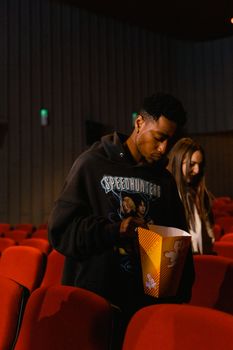 A man in a black hoodie holding popcorn inside a movie theater with red chairs.