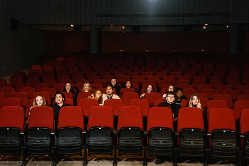 Un groupe de jeunes adultes est assis dans une salle de cinéma sombre, clairsemés sur plusieurs rangées de sièges rouges, regardant vers l'avant.