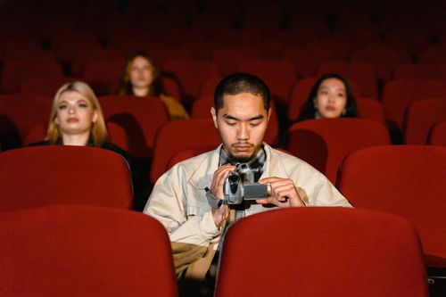 Free Man using a video camera in a dimly lit movie theater. Red seats surround him. Stock Photo