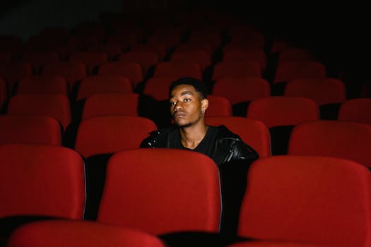 A lone man sits in an empty theater with red seats, looking contemplative.