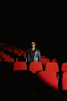 A fashionable man in sunglasses stands in a dark theater with striking red seats.