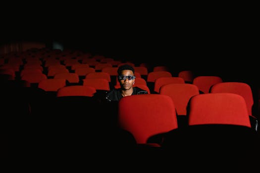 A solitary man wearing 3D glasses sits in an empty cinema hall with red seats.