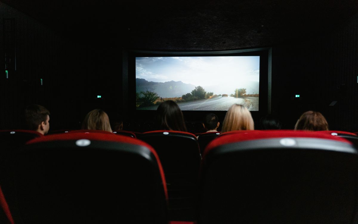 View from behind of people watching a movie in a cinema with red seats and a large screen.