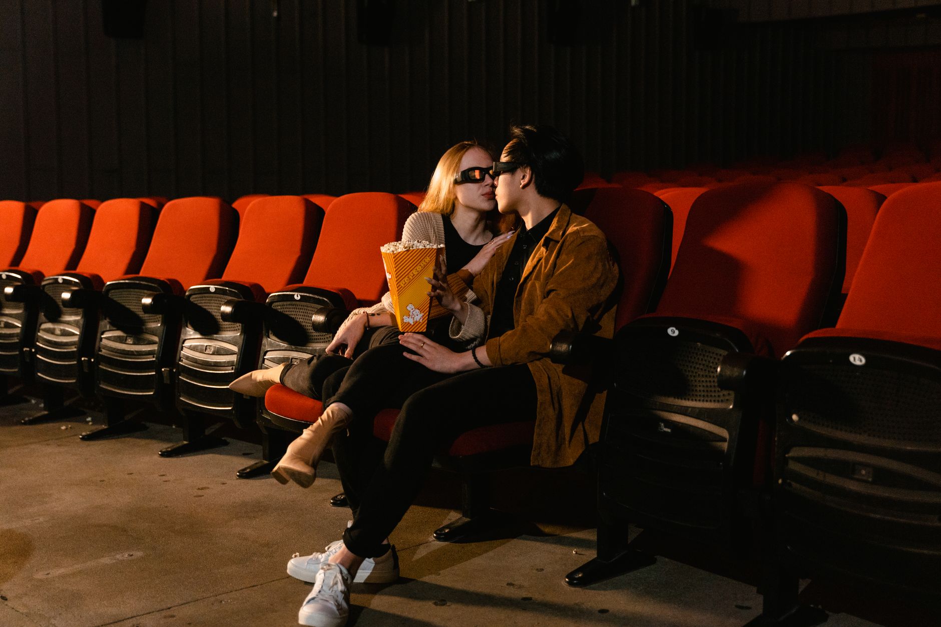 Couple wearing 3D glasses sharing popcorn in a cozy movie theater setting.