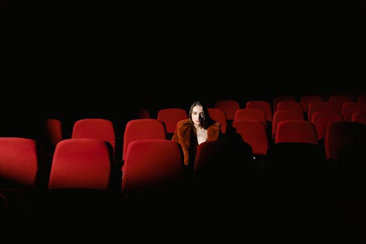 A woman sitting alone surrounded by empty red theater seats in a dark auditorium.
