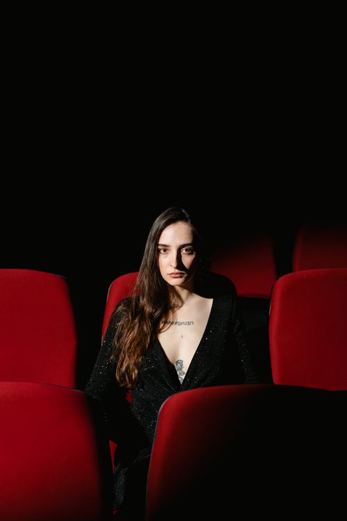 Free Fashionable woman in elegant attire sitting alone in a red-seated movie theater, dramatic lighting. Stock Photo