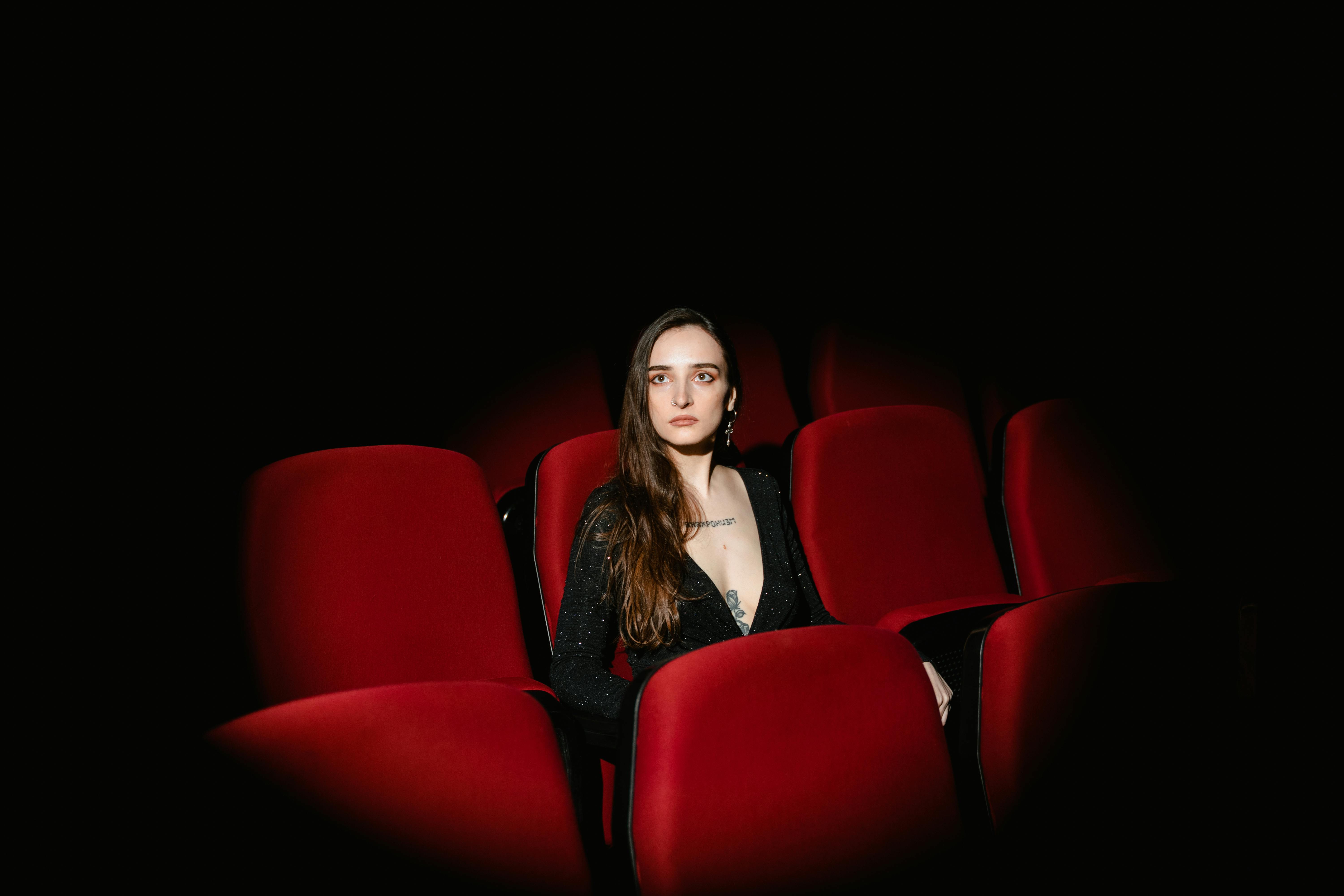 Free Stylish young woman sitting alone in a dimly lit movie theater. Stock Photo