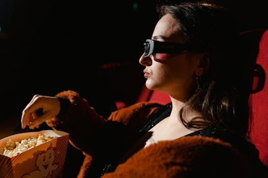 A woman wearing 3D glasses enjoys popcorn while watching a movie in a theater.