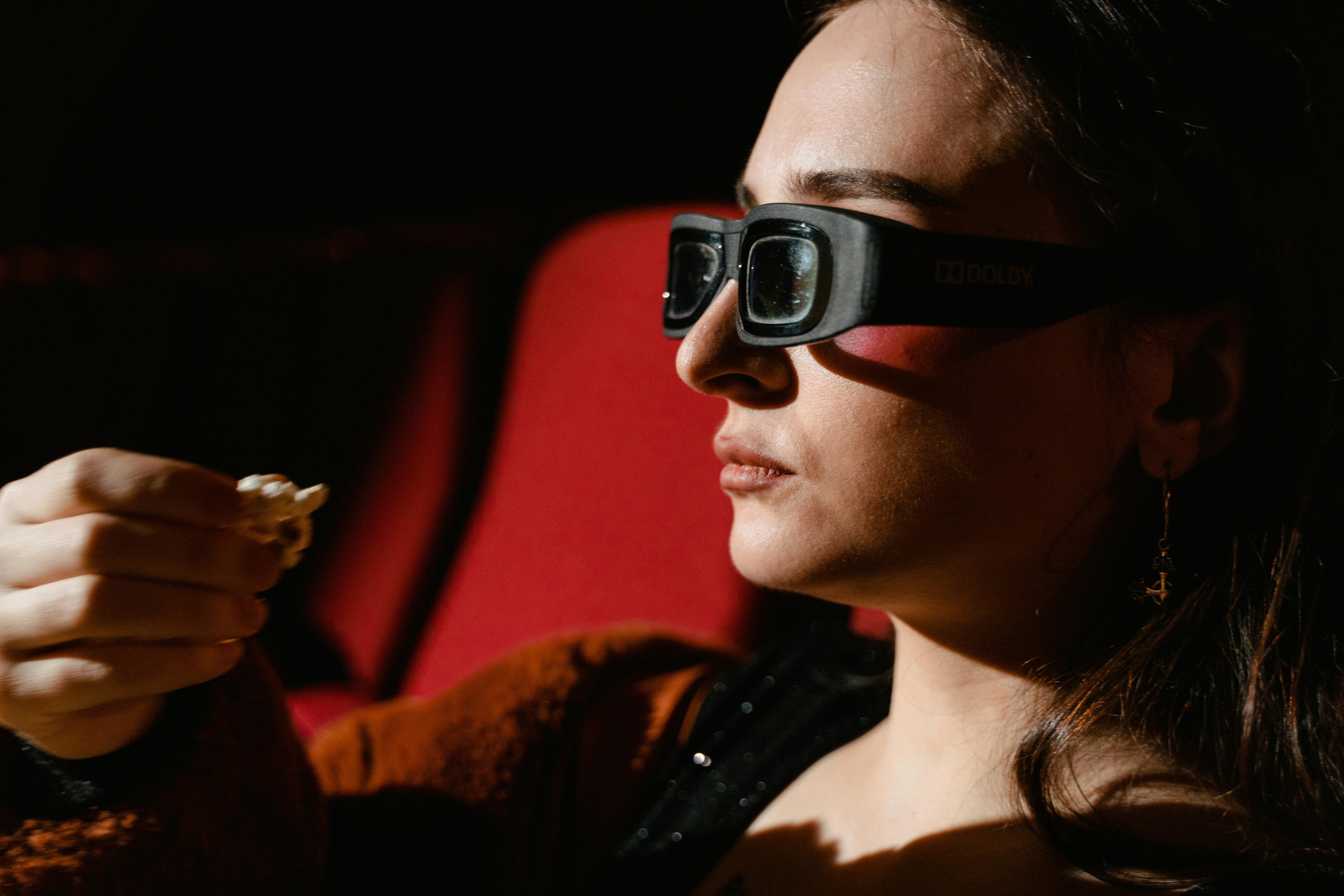 Free Side view of woman watching a movie in 3D glasses, holding popcorn in dimly lit cinema. Stock Photo