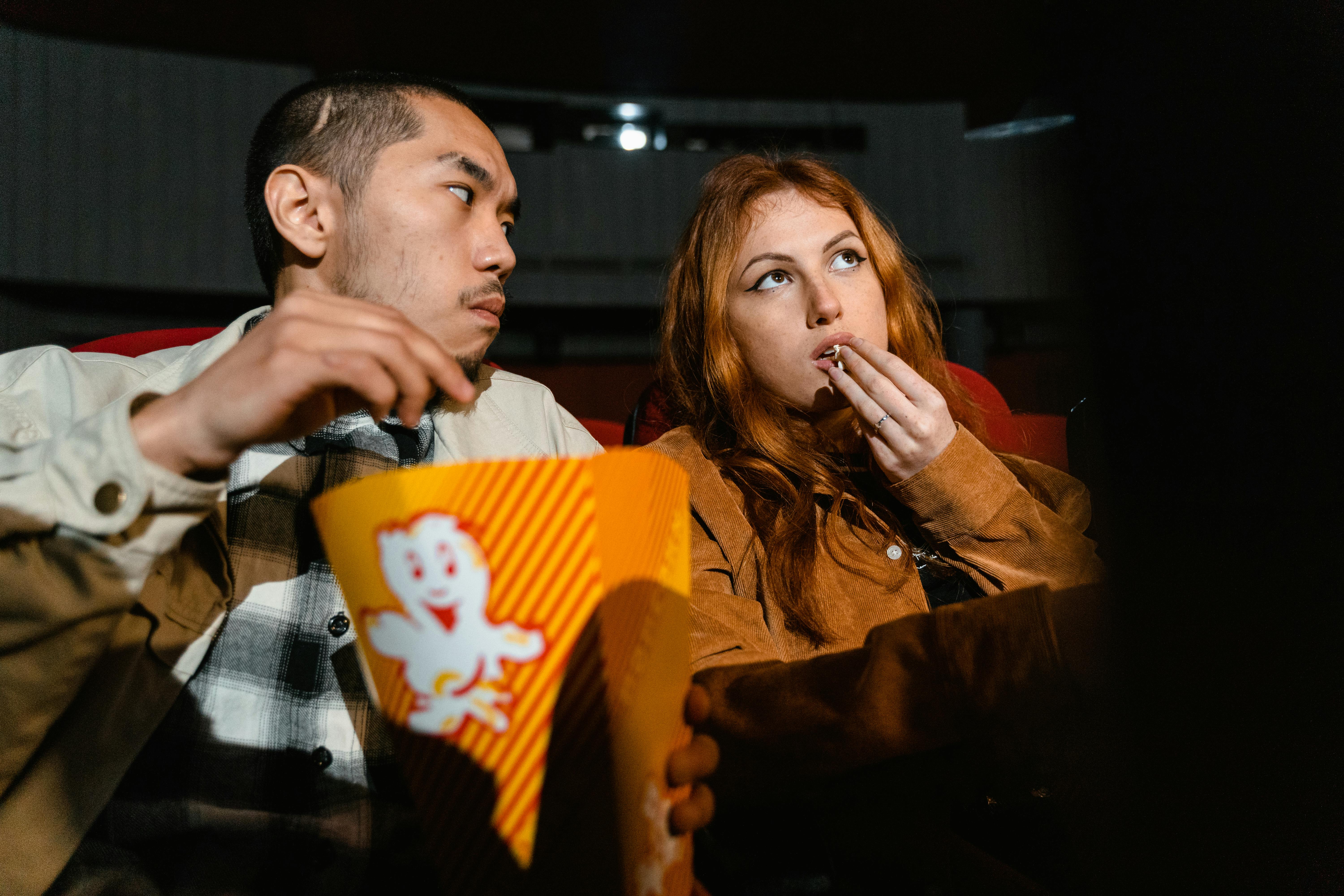 Free An Asian man and Caucasian woman enjoying popcorn together in a cozy movie theater. Stock Photo