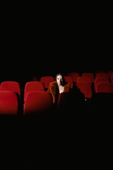 A woman seated alone in a dark, empty theater with red seats, creating a moody ambiance.