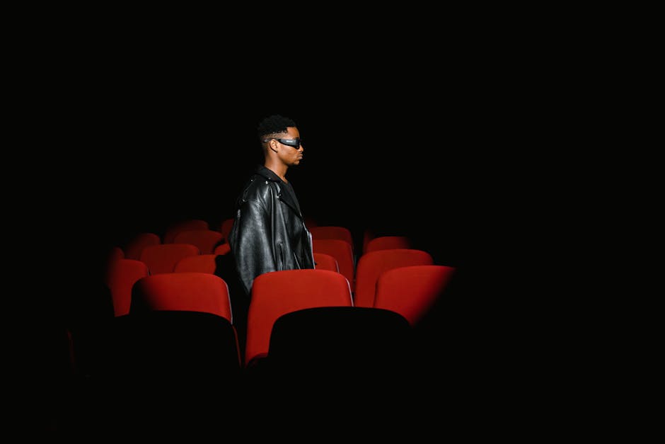 A person wearing a leather jacket and sunglasses in a dimly lit cinema with red seats.