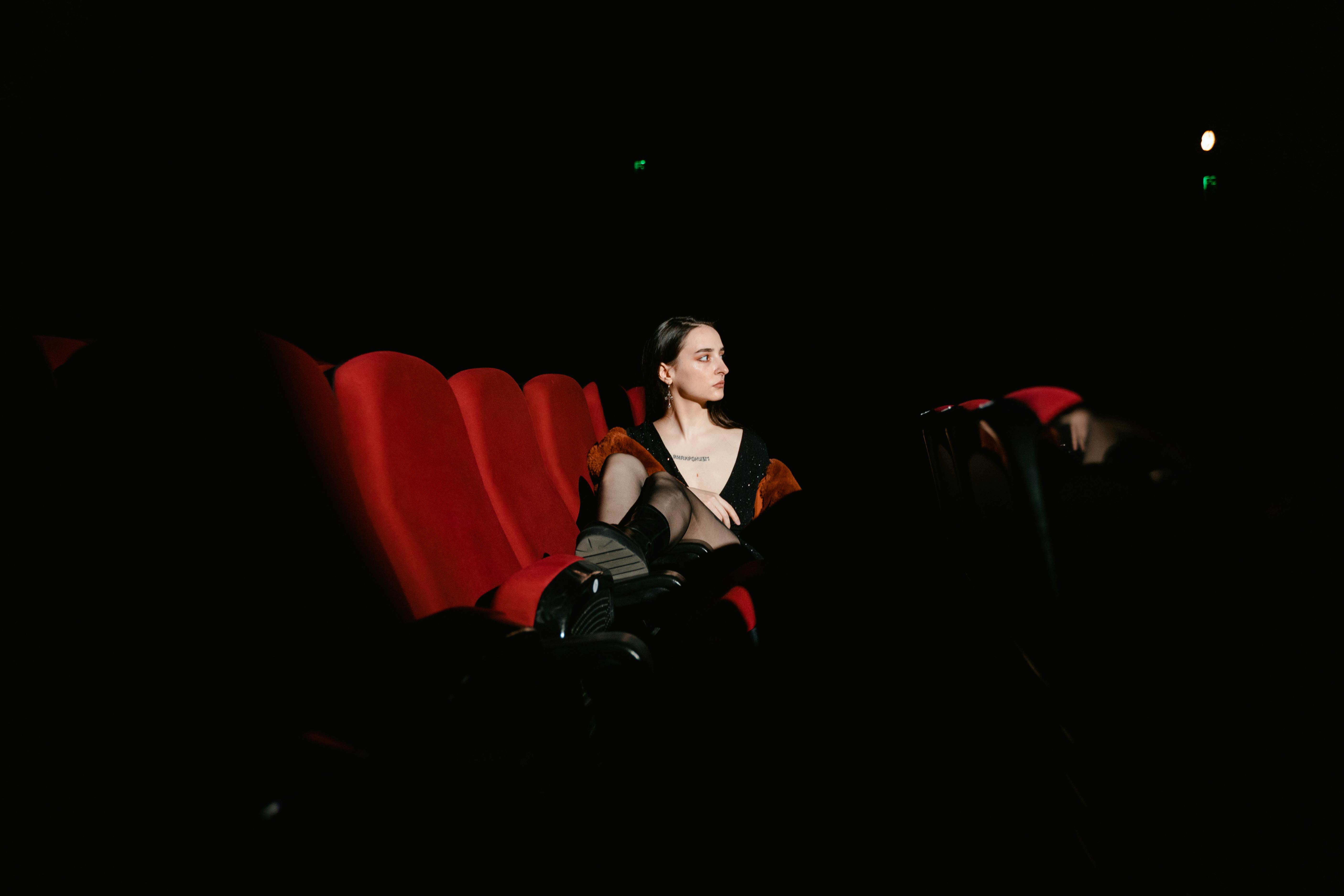 Free A lone woman sits in an empty theater, illuminated by soft light on red seats, creating a dramatic feel. Stock Photo