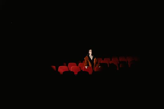 A woman in a fur coat sits alone in a dimly lit theater, surrounded by empty red seats.
