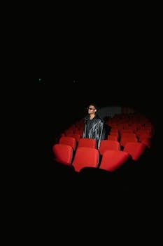 A man in a leather jacket sits alone in a dimly lit cinema with red seats.