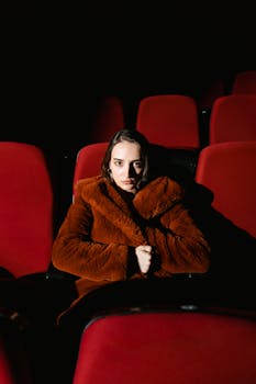 A woman in a fur coat sits alone in a red-seat cinema, looking pensive.