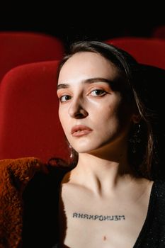 Close-up of a woman with a tattoo seated against a red background in dramatic lighting.