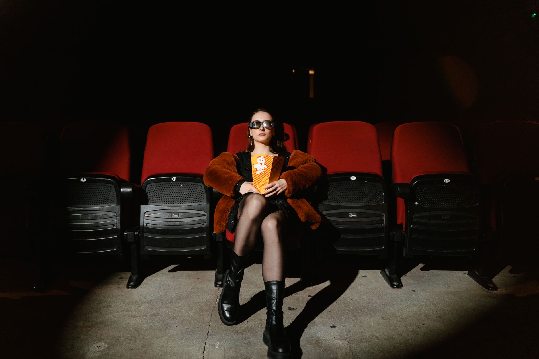 A stylish woman sits alone in a dark cinema, enjoying popcorn during a movie.