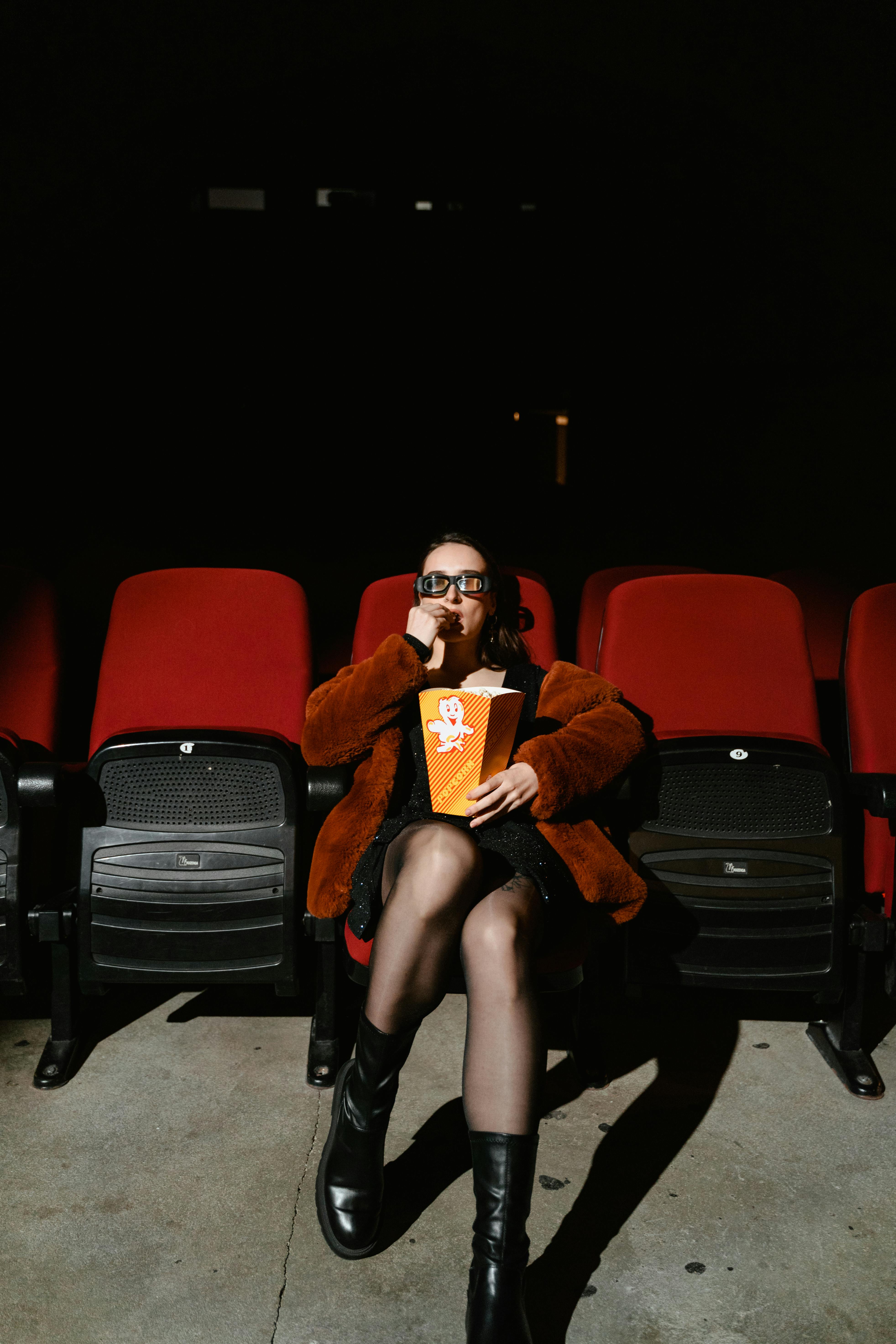 Free Woman alone in a theater, wearing 3D glasses, enjoying popcorn. Stock Photo