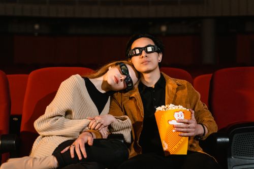 Free A cozy couple wearing 3D glasses, sharing popcorn in a cinema setting. Stock Photo