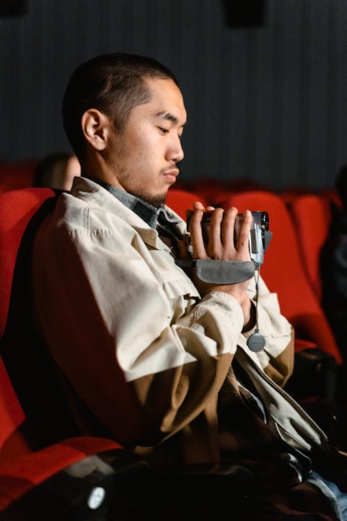 A Man in Beige Jacket Sitting while Filming Using a Video Camera