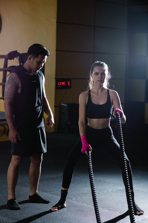 Full body Latin American personal instructor helping active female in activewear exercising with battle ropes during intense training in gym