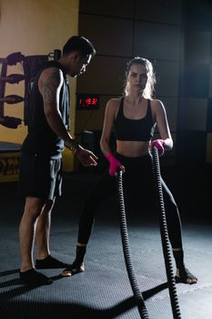 Female athlete training with battle ropes guided by her trainer in a gym.
