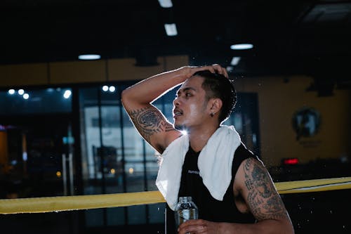 Free Side view of tired tattooed Latin American male in sportswear with white towel around neck standing in gym with bottle of water during workout Stock Photo