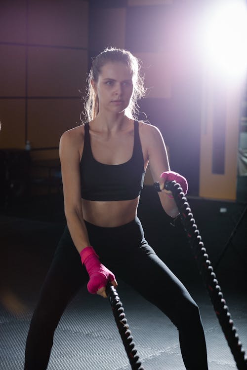 Determined sporty female in black activewear practicing with heavy battle ropes during intense training in modern gym with glowing light