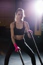 Determined sporty female in black activewear practicing with heavy battle ropes during intense training in modern gym with glowing light