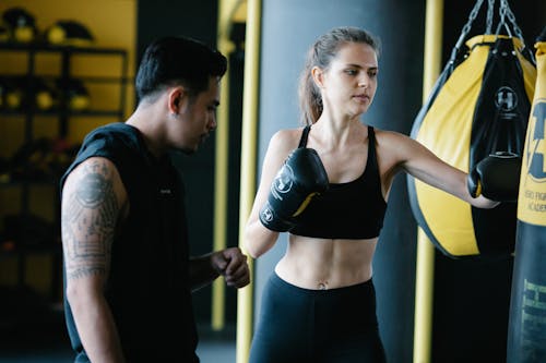 Muscular sportive female boxer practicing punches with heavy boxing bag during training with personal Latin American coach in modern gym