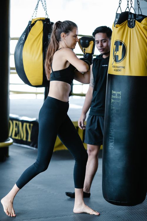 Free Sportive female in black activewear punching boxing bag during intense workout with Latin American personal instructor in light modern gym Stock Photo