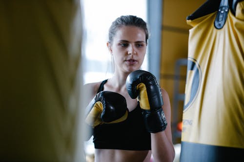 Sportive female in boxing gloves and black top standing near heavy punching bags during intense workout in light modern gym