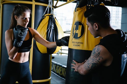 Sportive female boxer in gloves hitting heavy punching bag during intense boxing workout with personal Latin American trainer in gym