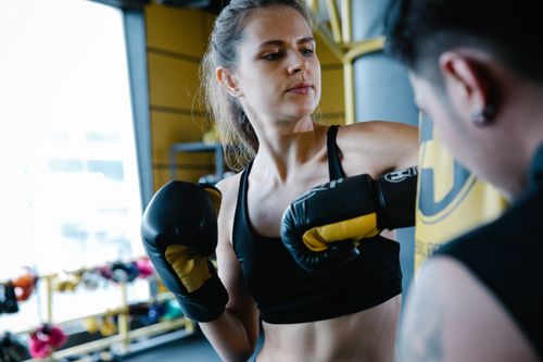 Free Determined female boxer in sportswear hitting heavy punching bag near anonymous personal instructor during workout in gym with special equipment Stock Photo