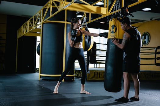 Full body of sportive female fighter punching boxing bag during workout with tattooed personal Latin American instructor in modern gym