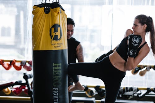 Strong female kickboxer in gloves kicking heavy punching bag near personal Latin American instructor during intense training in modern gym