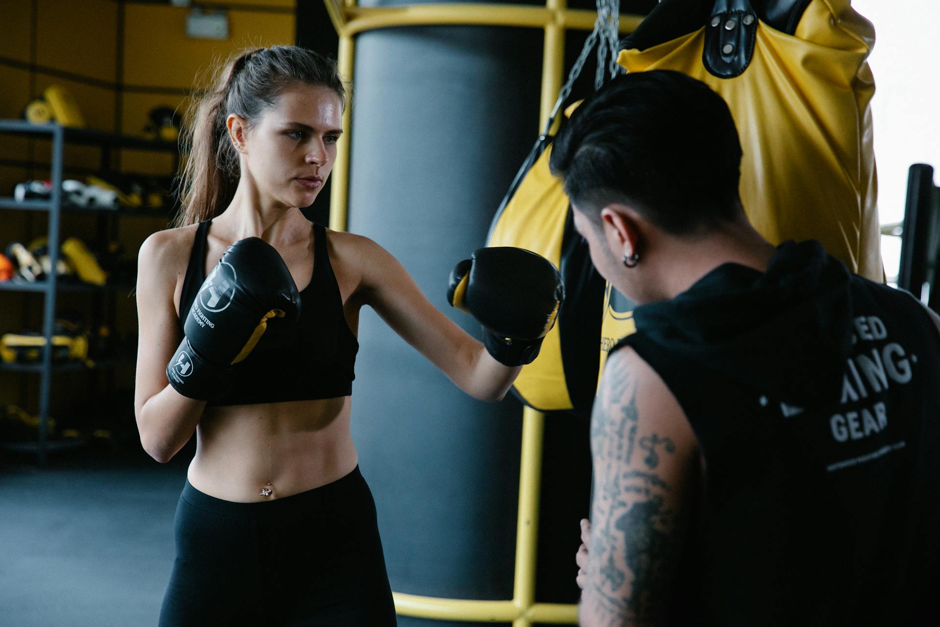 Anonymous personal trainer helping sportive female in gloves with punching technique near boxing bag during workout in light modern gym