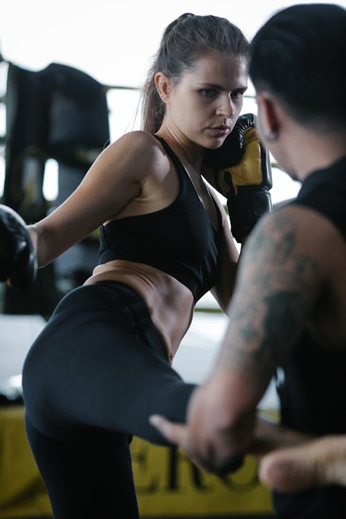 Determined young female fighter in sportswear and boxing gloves kicking male personal instructor during workout in gym