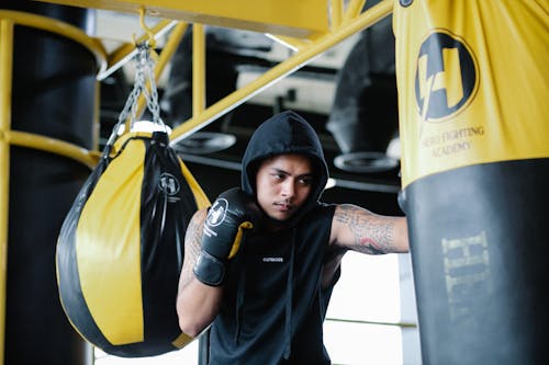 Free Focused young Latin American tattooed male fighter in hooded activewear and boxing gloves hitting punching bag during training in sports club Stock Photo