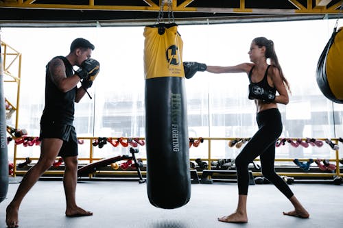 Side view of strong young female and male boxers in sportswear and protective gloves practicing punches on hanging heavy bag while training together in gym